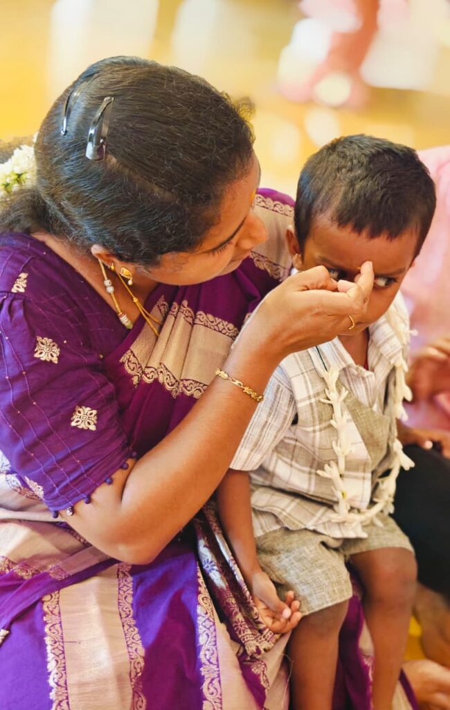 A woman in a purple saree places a mark on the forehead of a boy wearing a jasmine garland.