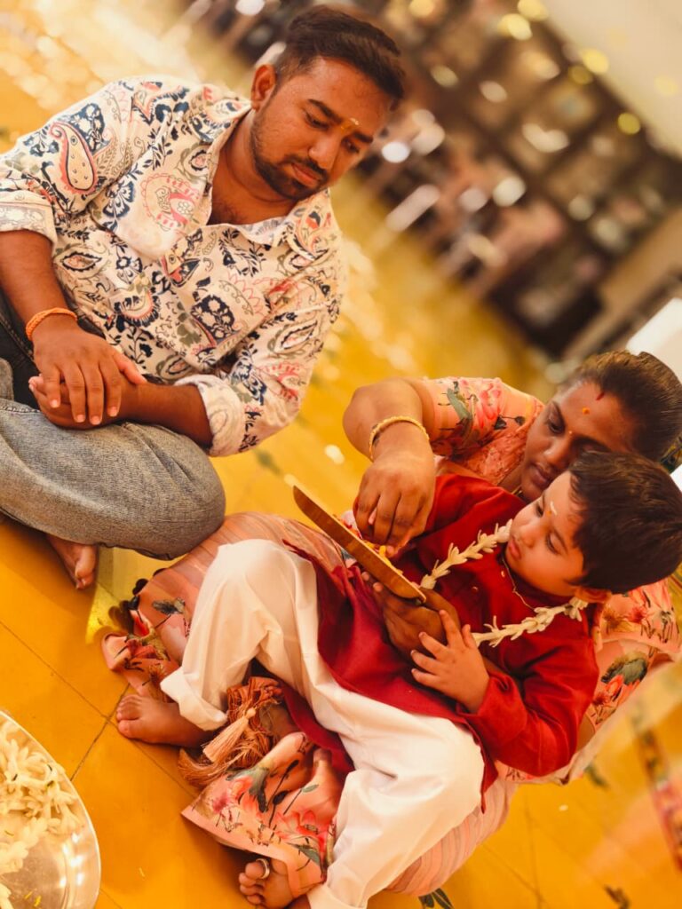 A child in a red kurta and garland traces in a tray held by a woman, with a man seated beside them.
