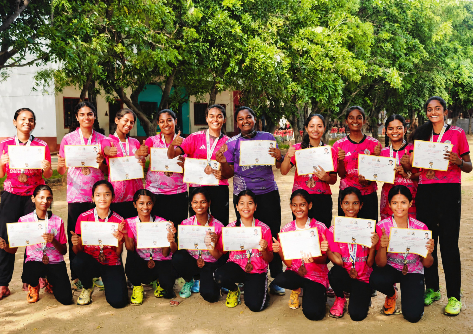 Girls in pink football jerseys pose under trees on a school ground, wearing medals and holding certificates.